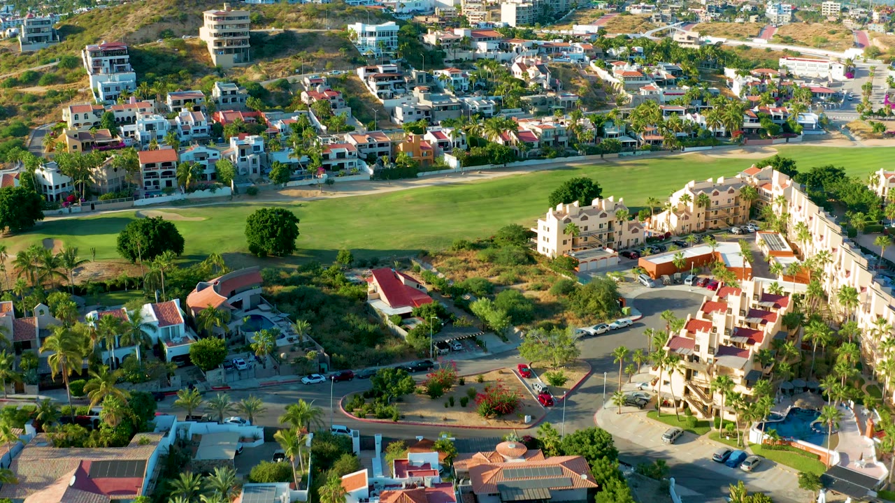 San Jose del Cabo aerial looking across a residential area where a green field spans the midground, hillside homes fill the background, and no visible people appear.