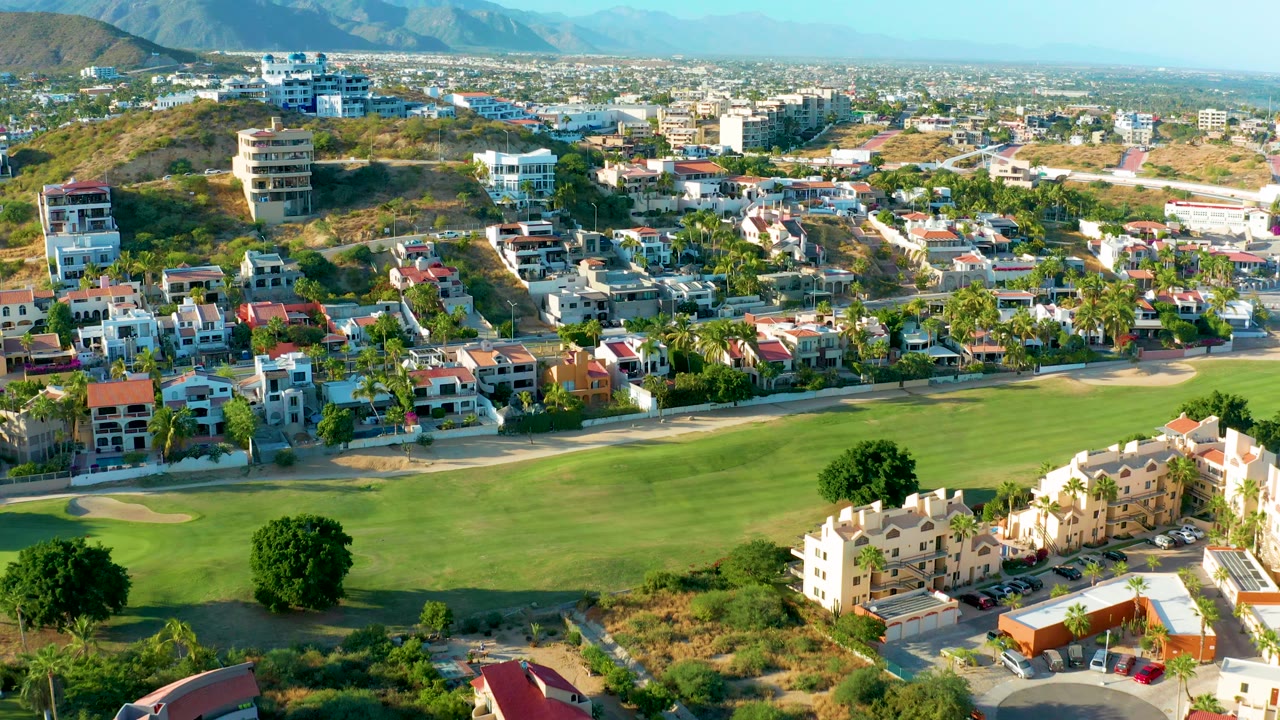 Wide drone view of a green golf fairway below residential streets and hills.