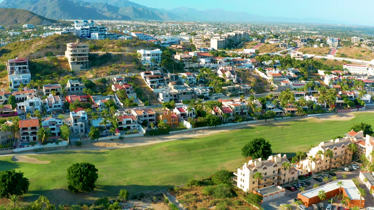 San Jose del Cabo aerial view with light-colored villas near the lower right, a broad golf course across the foreground, hillside homes, and mountains along the horizon, with no visible people.