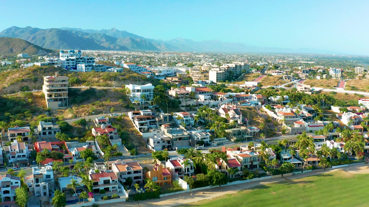 Opening aerial frame over a San Jose Del Cabo residential neighborhood with hillside homes, roads, palm trees, distant mountains, and no visible people.