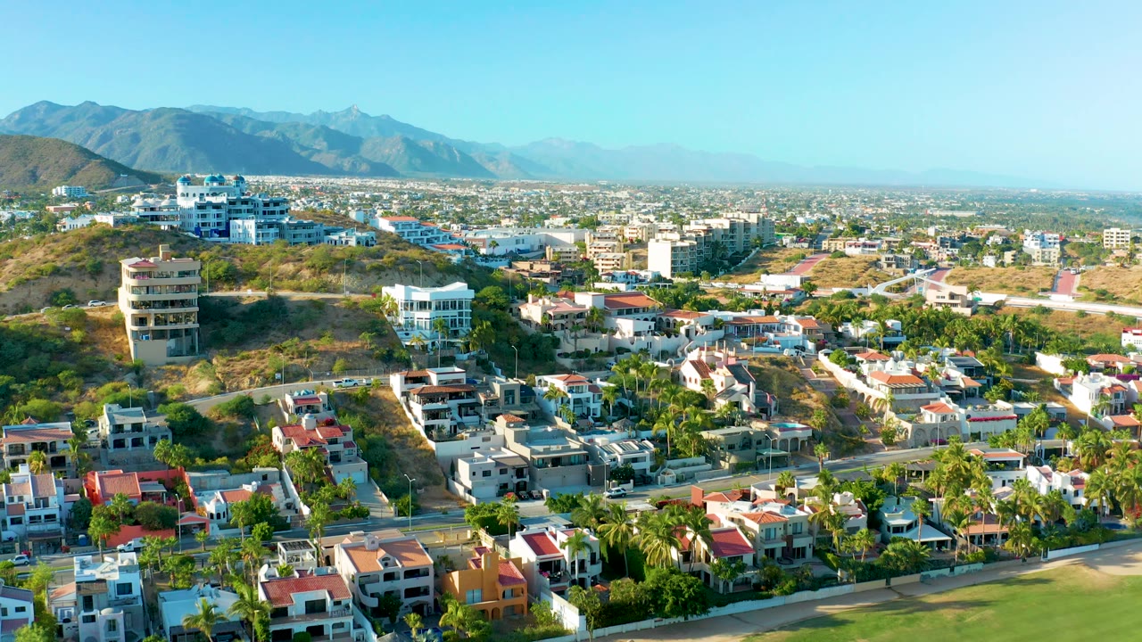 Third frame over San Jose Del Cabo with homes in the foreground, dry hillside lots, distant urban sprawl, mountains, clear sky, and no visible people.