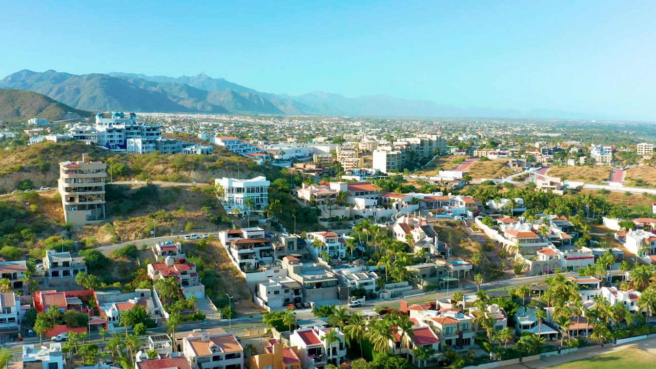 Drone view of houses along a road with palms, hillside buildings, and mountains in the background.