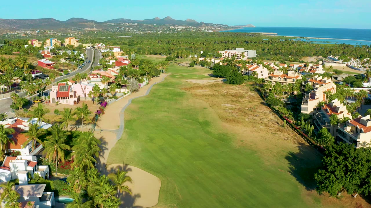 Wide aerial golf course view with fairway, sand trap, homes, palm trees, ocean, and mountains.