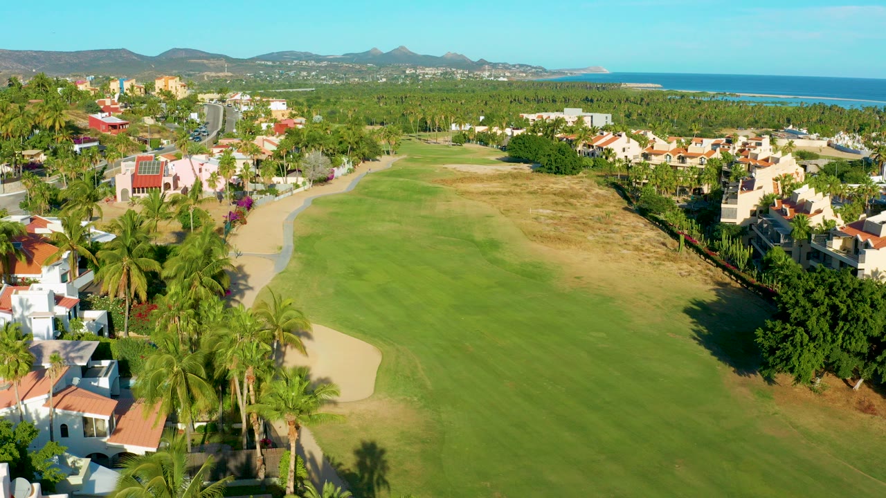 Aerial view in San Jose del Cabo showing a green golf fairway, sand trap, palm trees, homes, distant hills, ocean horizon, and no visible people.