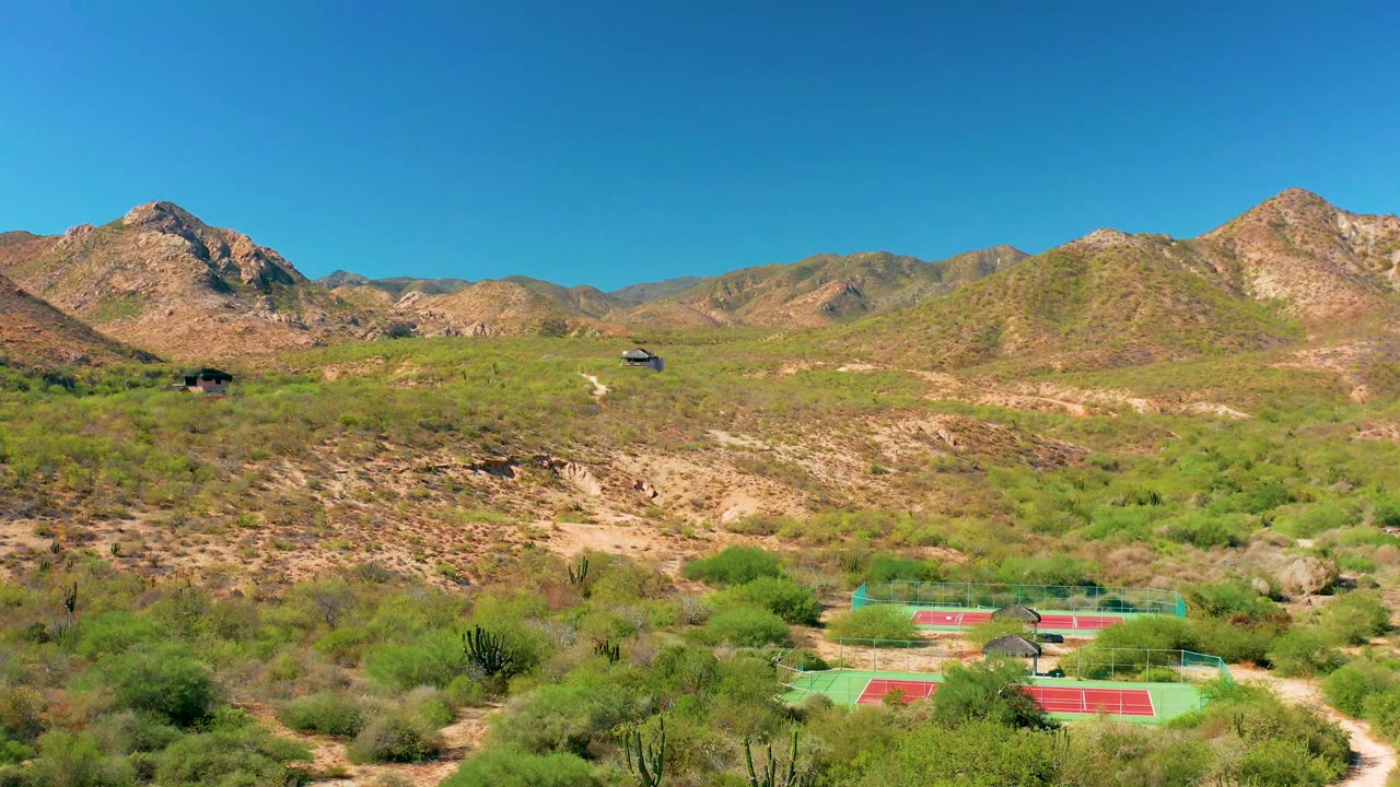 Aerial Cabo Pulmo view with the courts still near the lower edge, a pale dirt track curving through scrub, rocky hills across the middle distance, and no visible people.