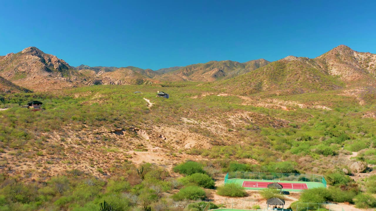 Aerial frame in Cabo Pulmo, Baja California Sur with scrub-covered hills, pale dirt tracks, small distant buildings, rocky mountain ridges, clear blue sky, and no visible people.