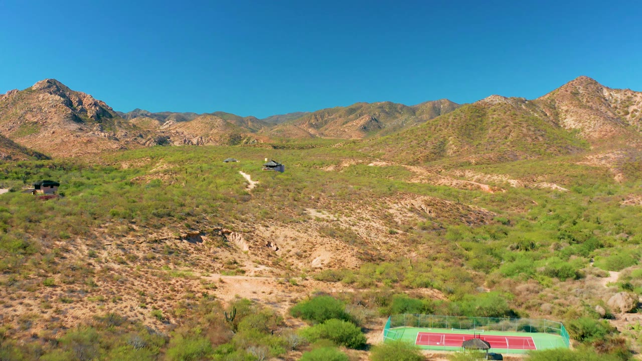 Aerial frame in Cabo Pulmo, Baja California Sur showing dry green-brown hills, a winding dirt path, distant small structures, rugged mountain ridges, blue sky, and no visible people.