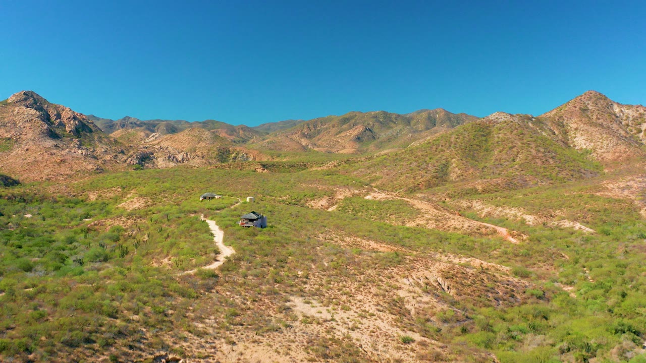 Wide aerial frame of Cabo Pulmo desert hills with two small vehicles on a pale dirt trail, rocky mountains across the horizon, clear blue sky, and no visible people.