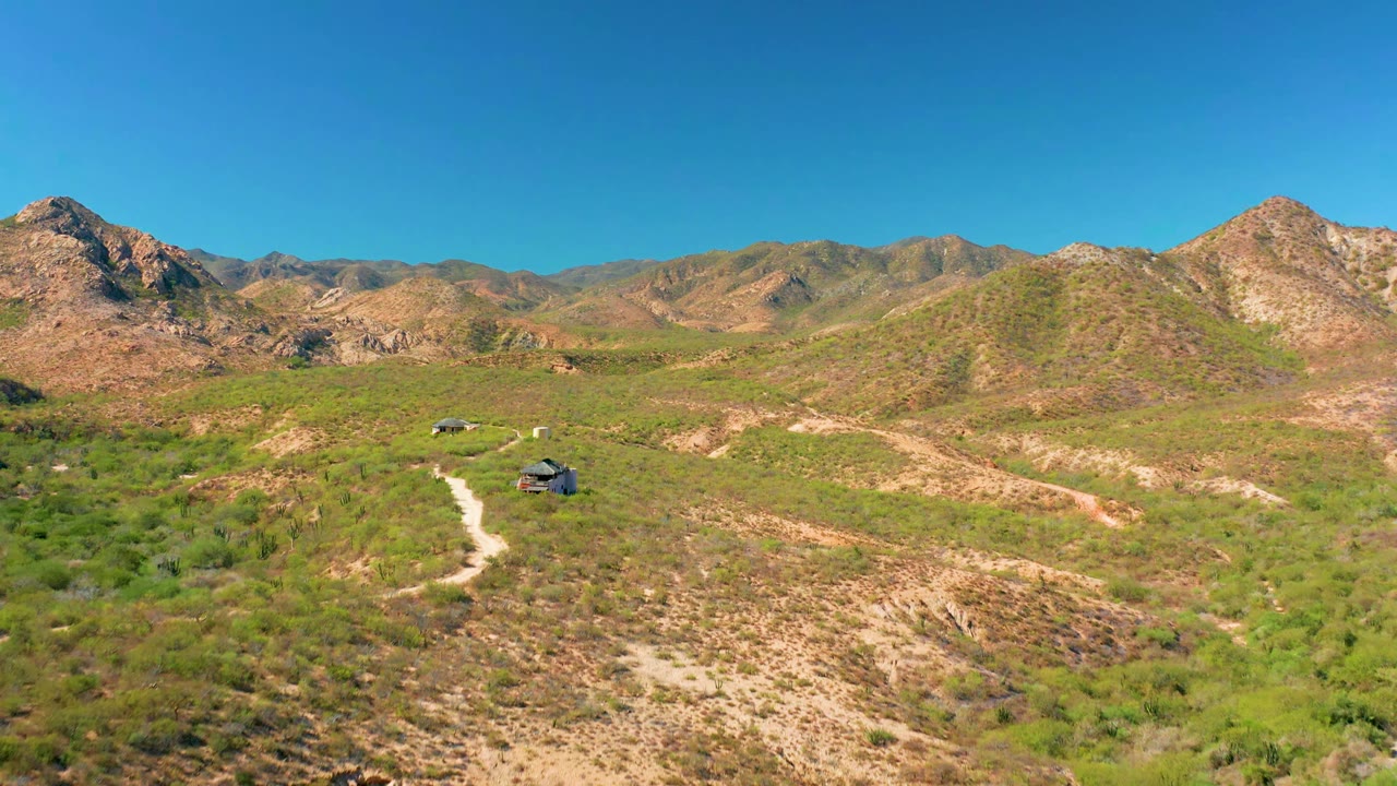 Aerial view in Cabo Pulmo with a curving dirt path, two vehicles separated along the trail, green desert vegetation, tan rocky slopes, and no visible people.