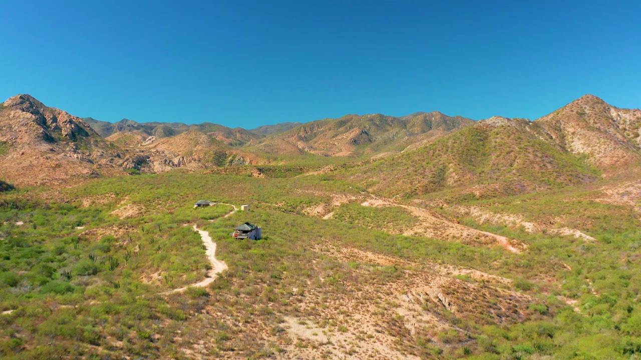 Cabo Pulmo aerial frame with two vehicles on a light dirt road, sparse green brush in the foreground, layered rocky hills behind, and no visible people.