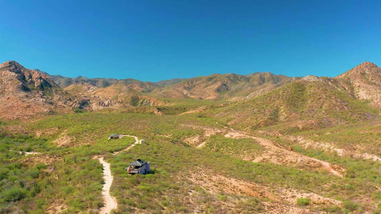 Cabo Pulmo aerial frame with the dirt road leading up from the lower left toward the SUV, scrub-covered hills, distant mountain ridges, clear sky, and no visible people.