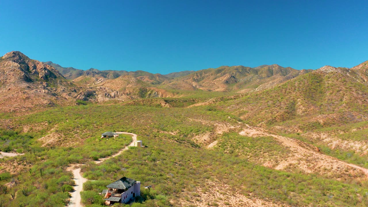 Cabo Pulmo, Baja California Sur aerial view opens on green scrub, pale dirt tracks, a small roofed structure at the lower edge, rocky hills, blue sky, and no visible people.