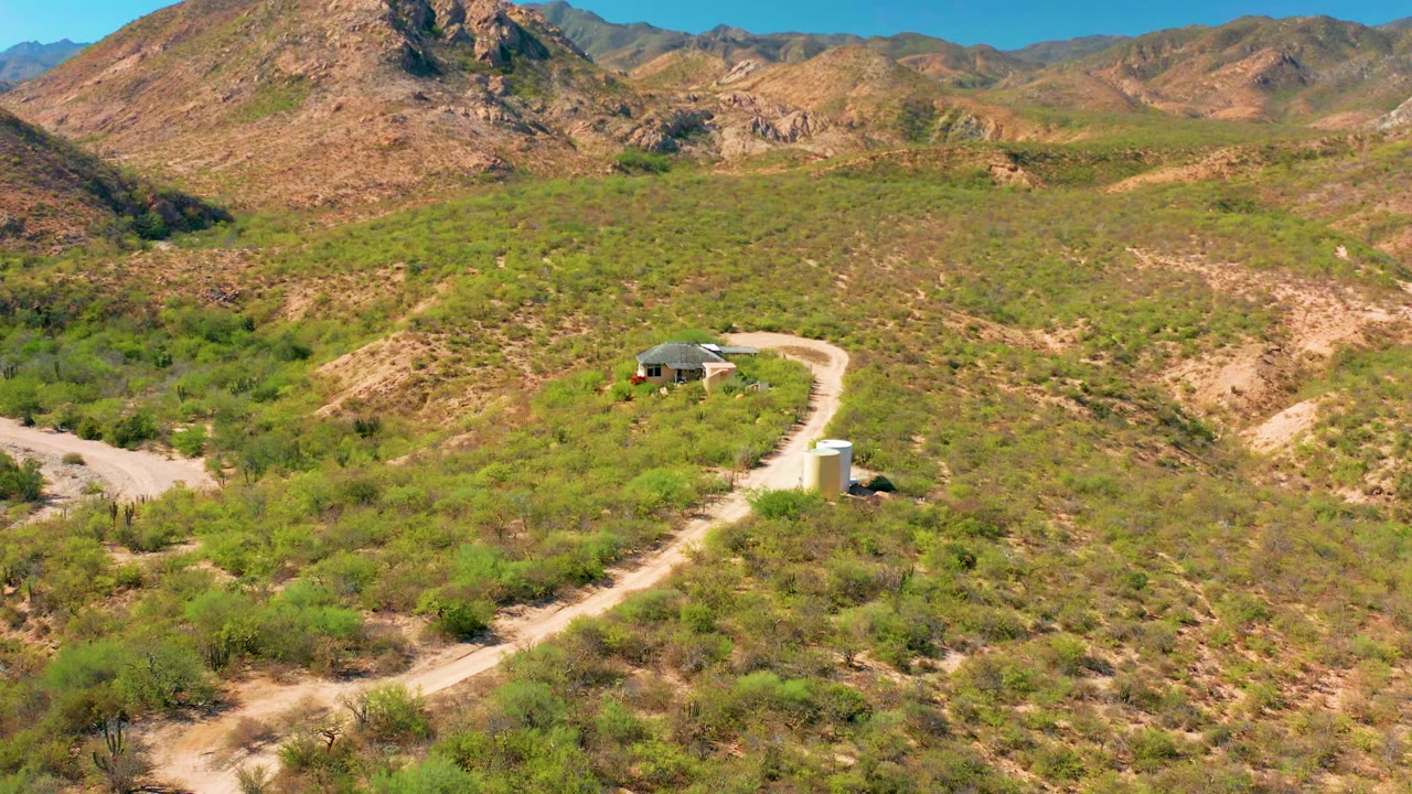 Aerial view in Cabo Pulmo, Baja California Sur showing a winding dirt road, sparse green desert vegetation, rocky hills, and a small structure near the road with no visible people.