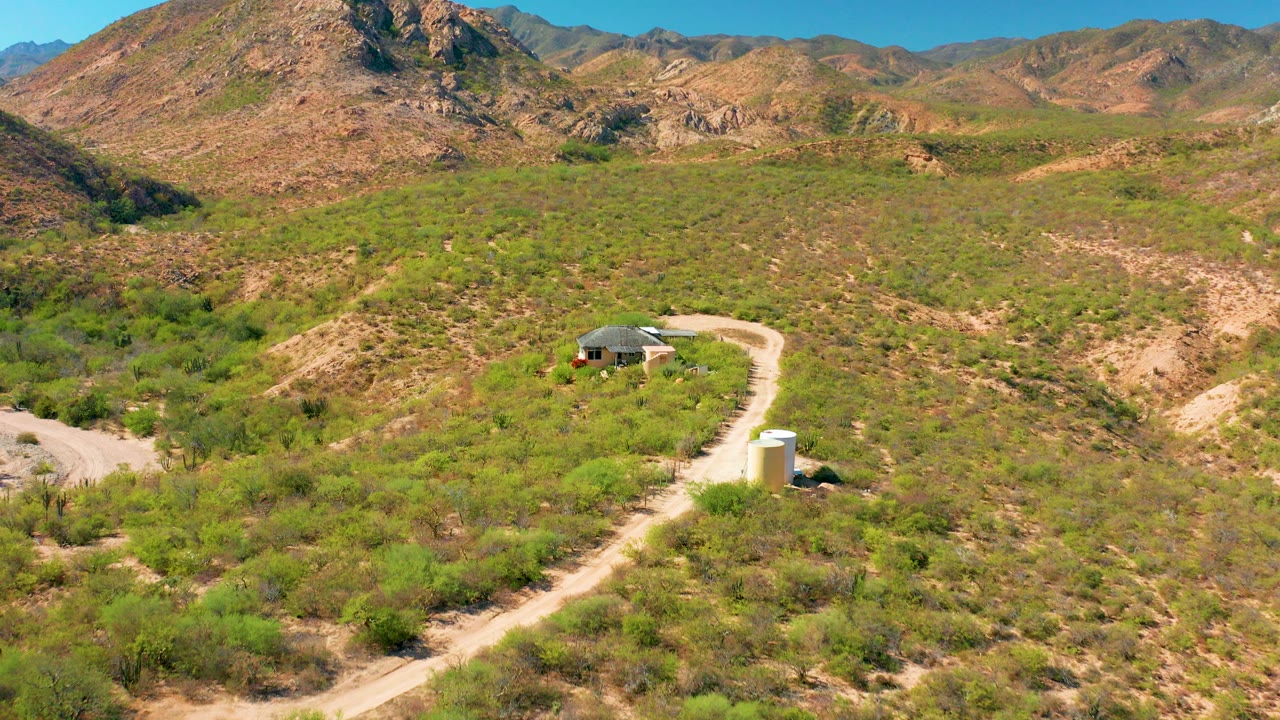 Aerial frame from Cabo Pulmo, Baja California Sur, showing a small rural house beside a curving dirt road, a white cylindrical structure in the foreground, green desert vegetation, and mountains in the distance.