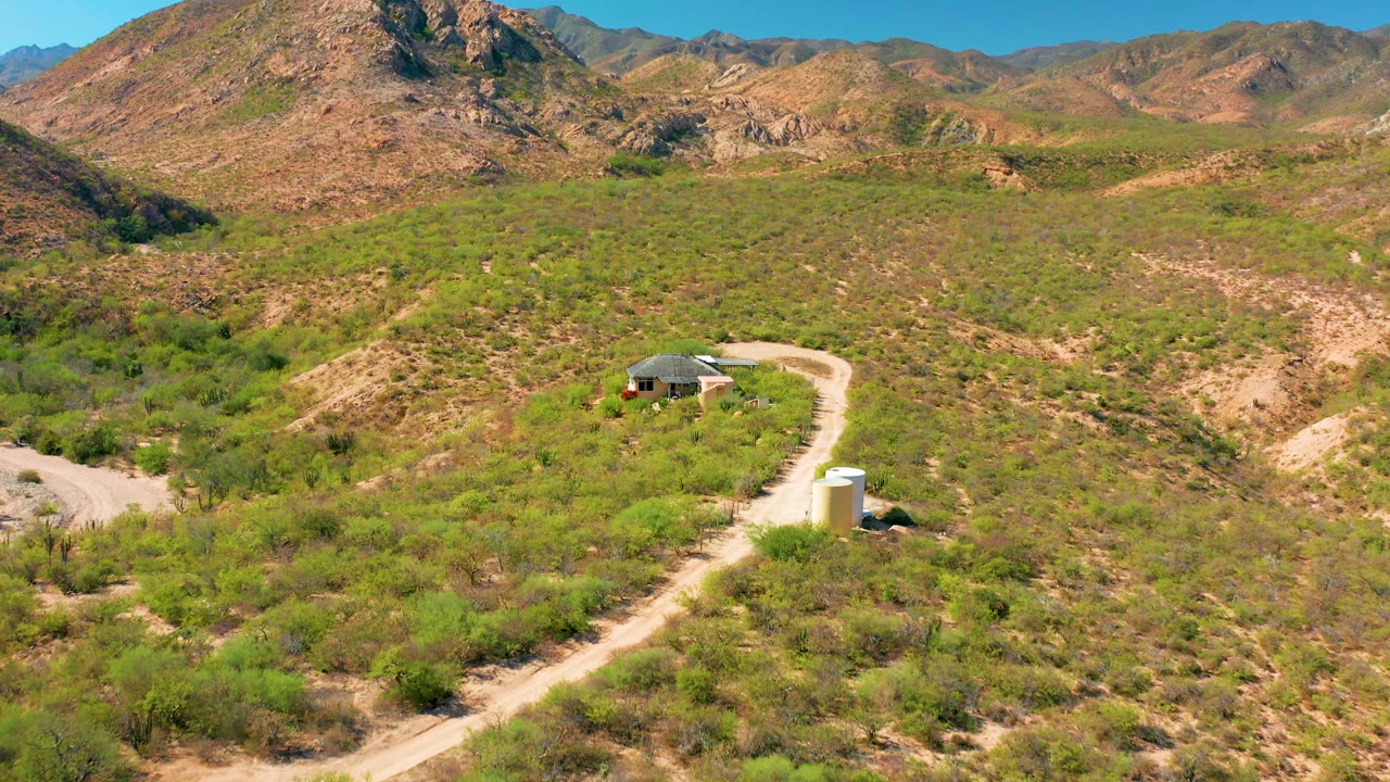 High aerial view of a dirt road leading past a white tank toward a small house in desert hills.
