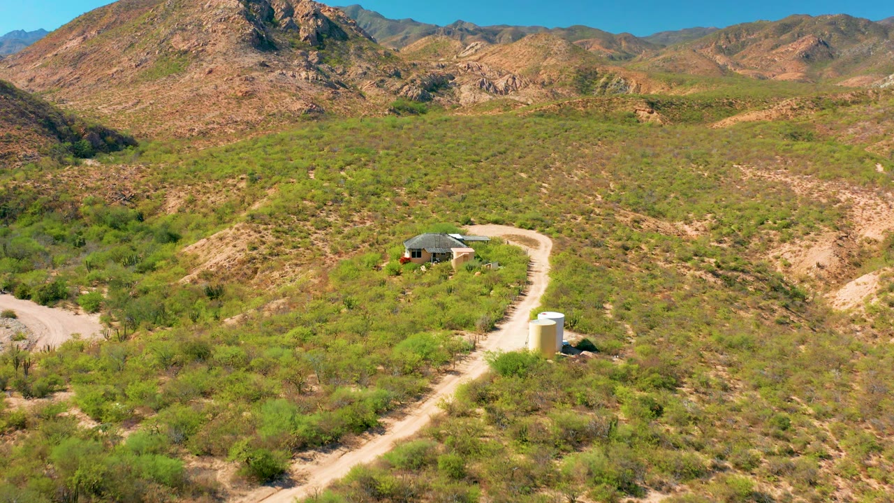 Aerial frame from Cabo Pulmo, Baja California Sur, showing the rural house near the left side of the road, the white cylindrical structure lower in frame, and layered desert hills behind it.