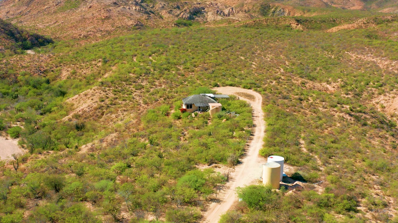 Aerial frame over Cabo Pulmo, Baja California Sur showing a small rural house, a pale winding dirt road, a white cylindrical tank, green-brown scrub hills, and no visible people.