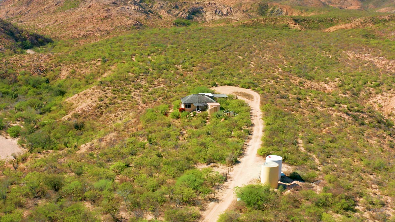 A slightly closer aerial view in Cabo Pulmo shows the dirt road curving past scrubby slopes toward a single-story house, with the white tank lower in frame and no visible people.