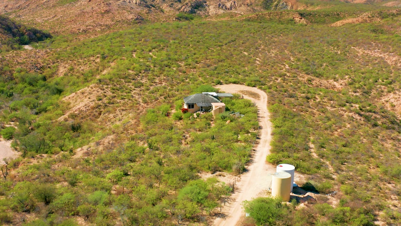 Aerial footage over Cabo Pulmo shows the house more centered near the winding road, with low green vegetation, brown rocky slopes, a white tank, and no visible people.
