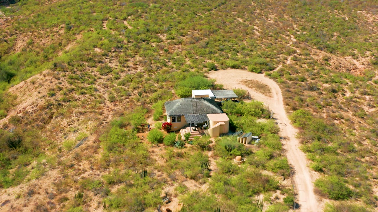 Aerial view in Cabo Pulmo, Baja California Sur, where the small house sits lower in the frame beside a bright dirt road, surrounded by scrub vegetation and dry ridges, with no visible people.