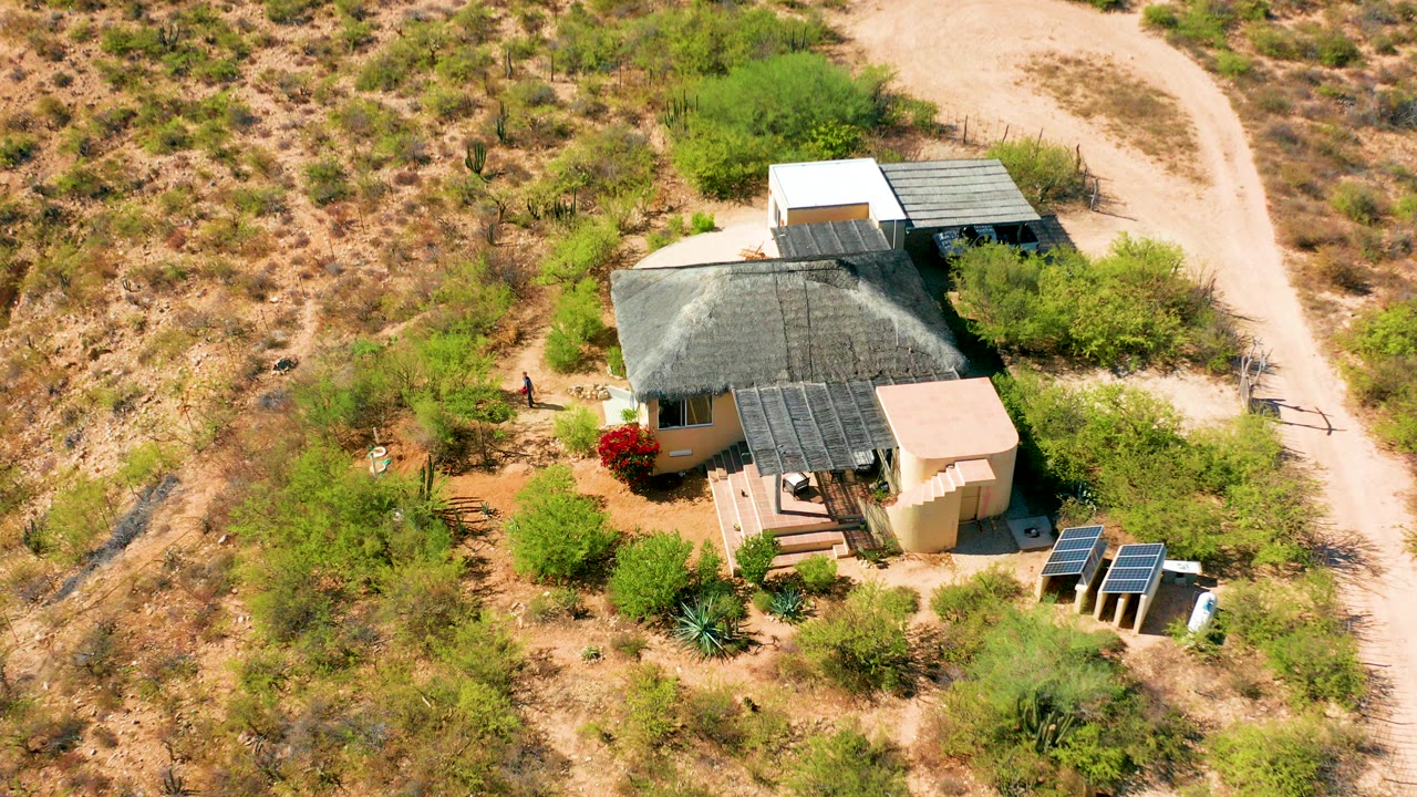 A wide aerial view in Cabo Pulmo, Baja California Sur shows a beige desert house with solar panels, sparse shrubs, a pale dirt road, and a small person near the left side of the property.