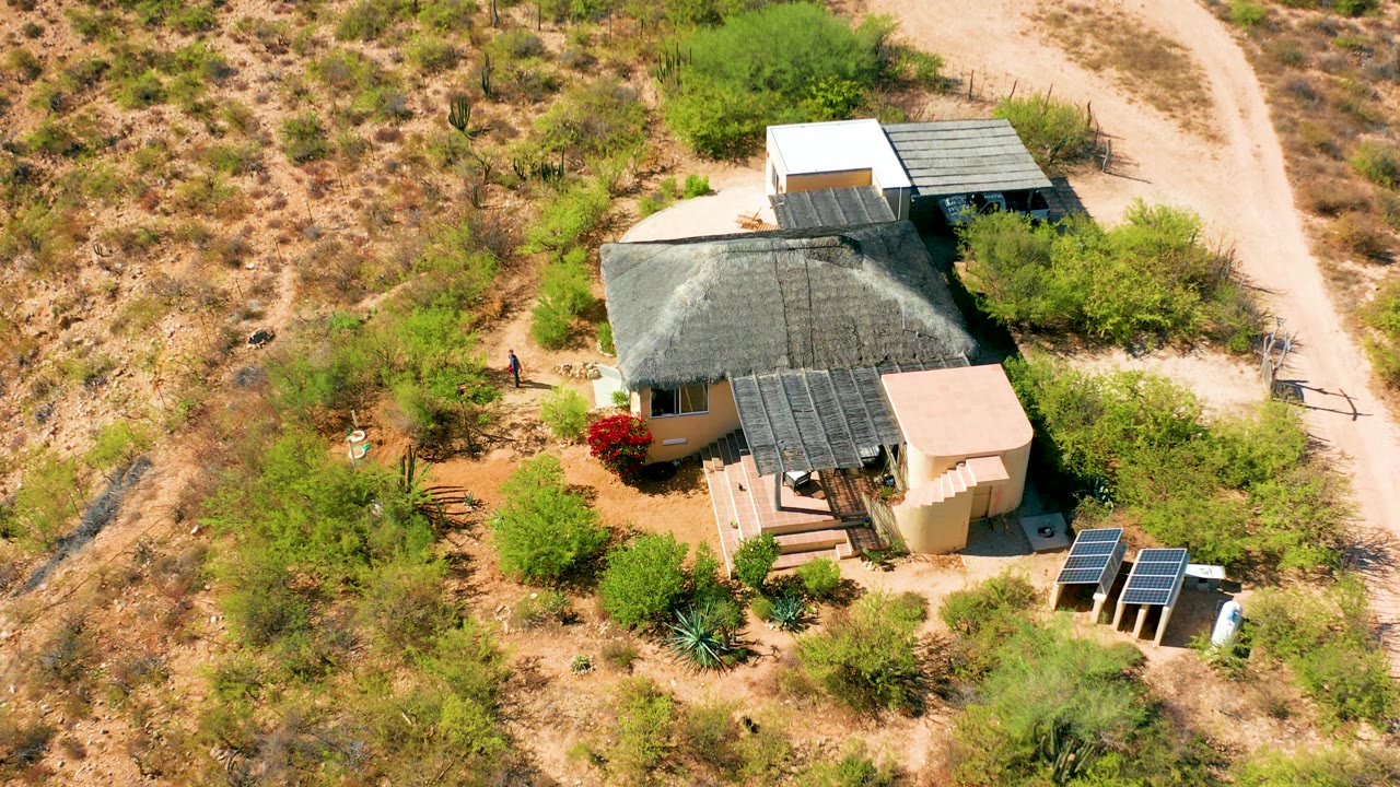 A closer overhead frame shows the Cabo Pulmo home centered below the camera, with solar panels at the lower right, steps at the front, a dirt road along the right edge, and a small person left of the house.