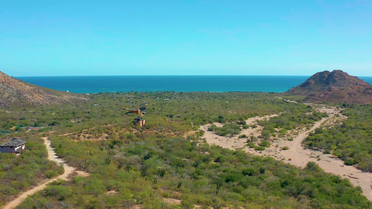 Cabo Pulmo aerial frame with the vehicle slightly farther along the dirt track, surrounded by scrub vegetation, branching sandy paths, hills, ocean, clear blue sky, and no visible people.