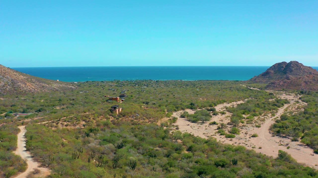 At 0:00, a wide aerial view in Cabo Pulmo shows sandy dirt tracks crossing green desert scrub toward low hills and a blue ocean horizon, with no visible people.