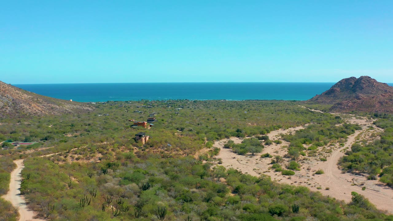 At 0:02, the Cabo Pulmo aerial angle shows the main sandy track splitting through desert vegetation, with hills on both edges and calm blue ocean beyond, with no visible people.