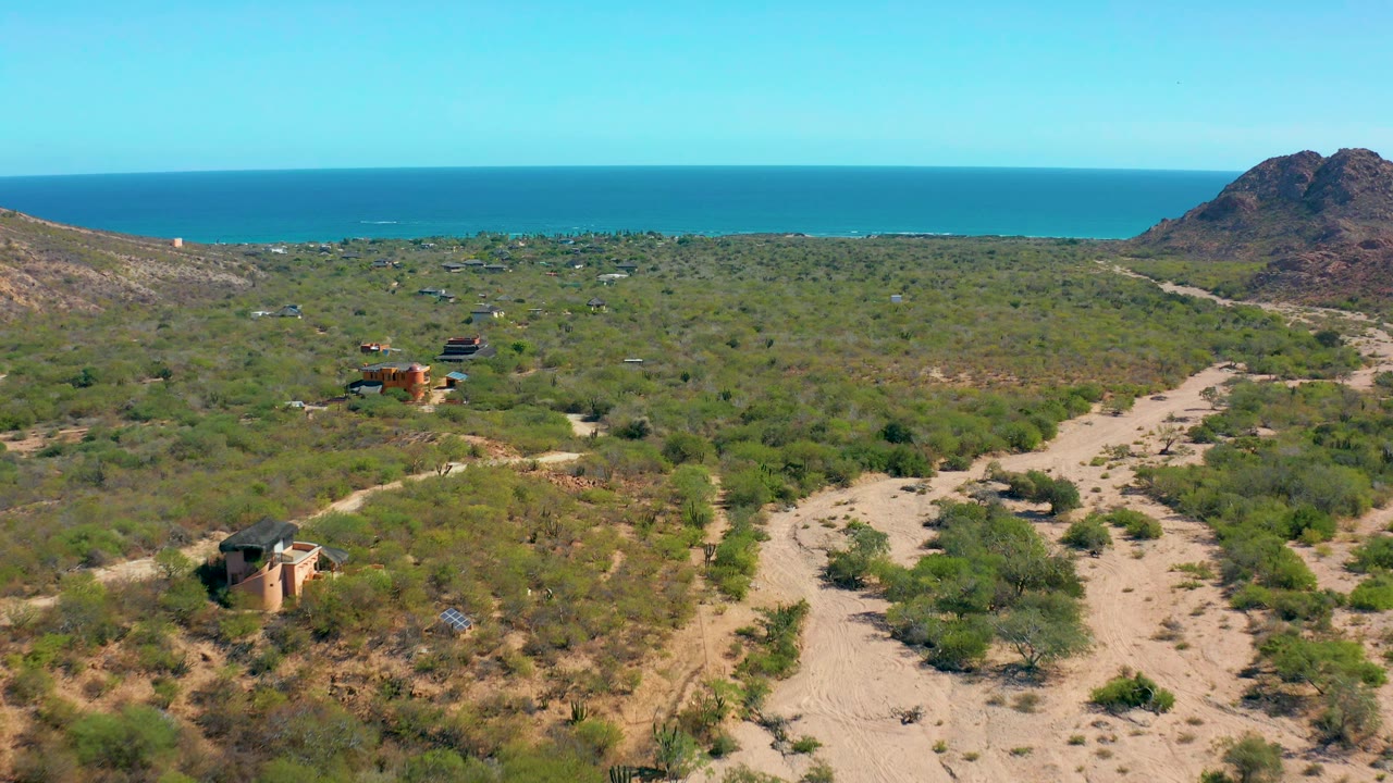 High aerial view over dry shrubland with a sandy wash, small buildings, ocean, and rocky hill.