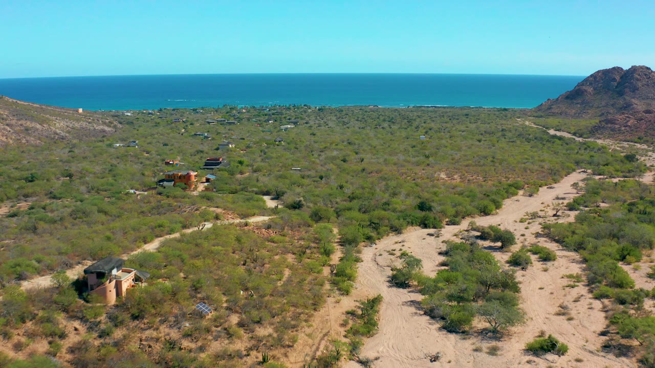Aerial frame in Cabo Pulmo showing winding sandy tracks through dense shrubs, several scattered structures, a rocky hill at the coast, the blue ocean horizon, and no visible people.