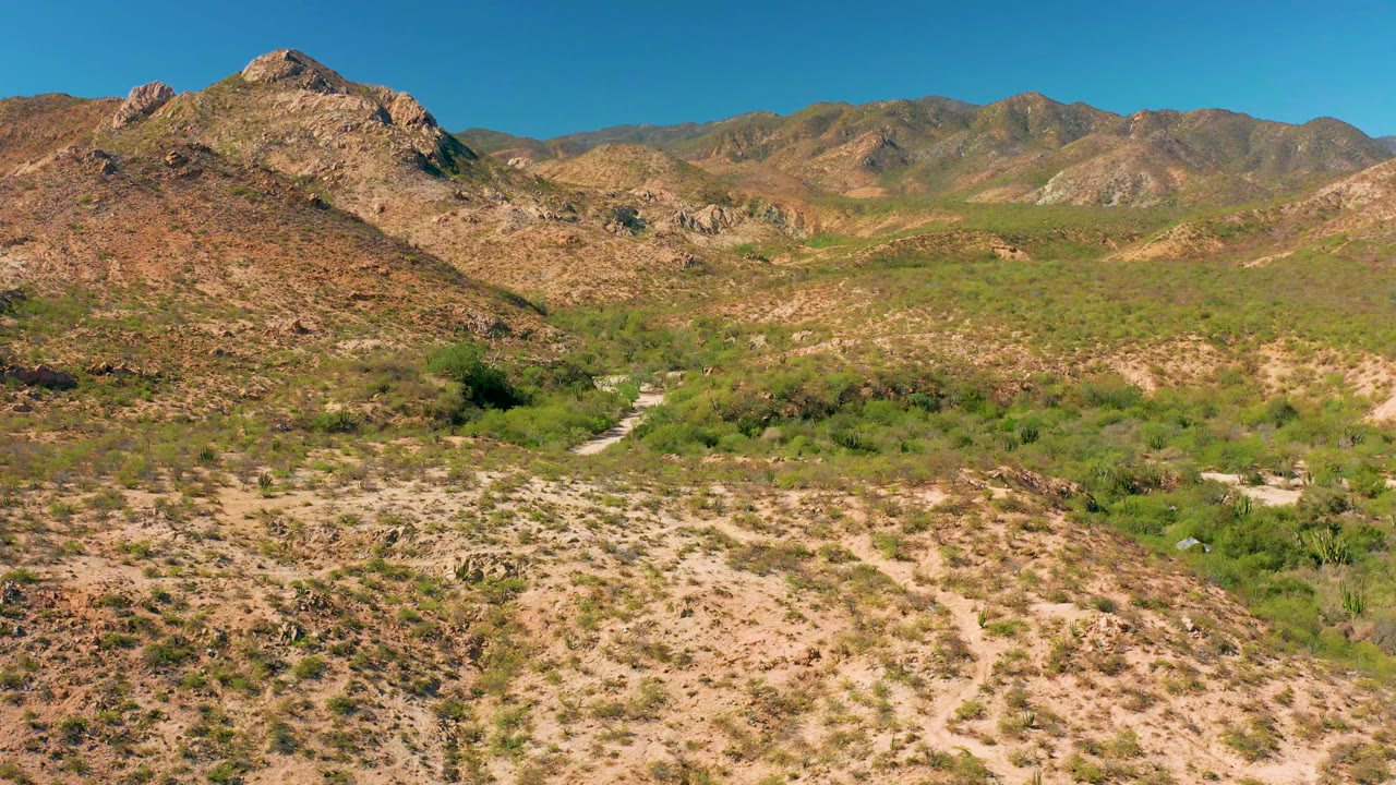 Cabo Pulmo aerial frame showing tan rocky slopes, low green desert vegetation, a pale wash through the foreground, distant mountain ridges, and no visible people.