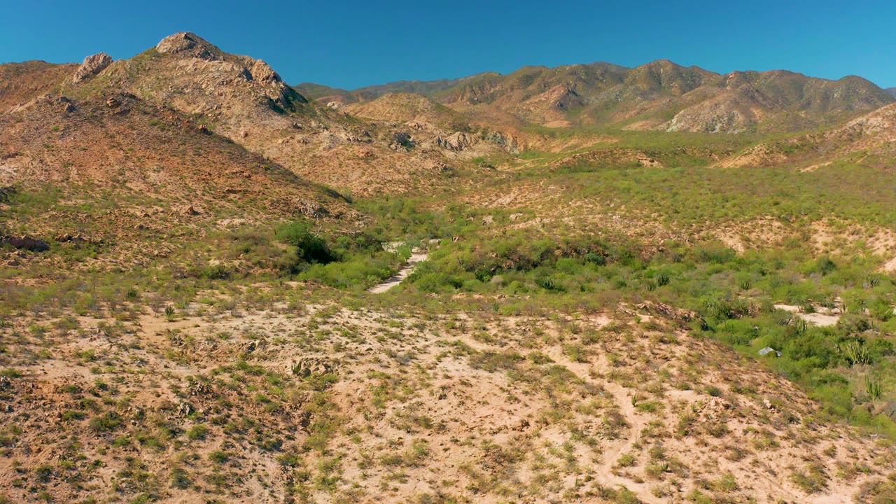 Cabo Pulmo aerial landscape with scrubby green growth running through the low ground, rocky desert hills on both sides, distant mountains, bright daylight, and no visible people.