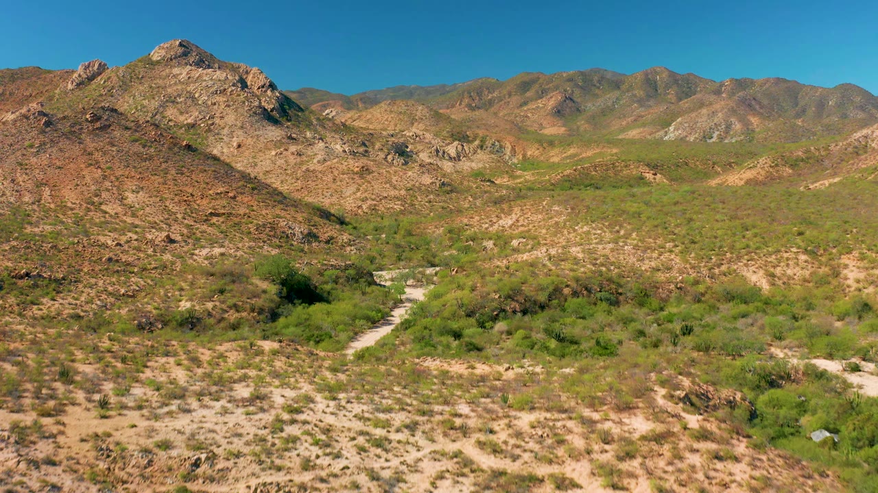 Aerial frame in Cabo Pulmo showing the central rocky hill slightly closer, scrub-covered desert slopes, the pale road bending through the lower valley, distant ridges, and no visible people.