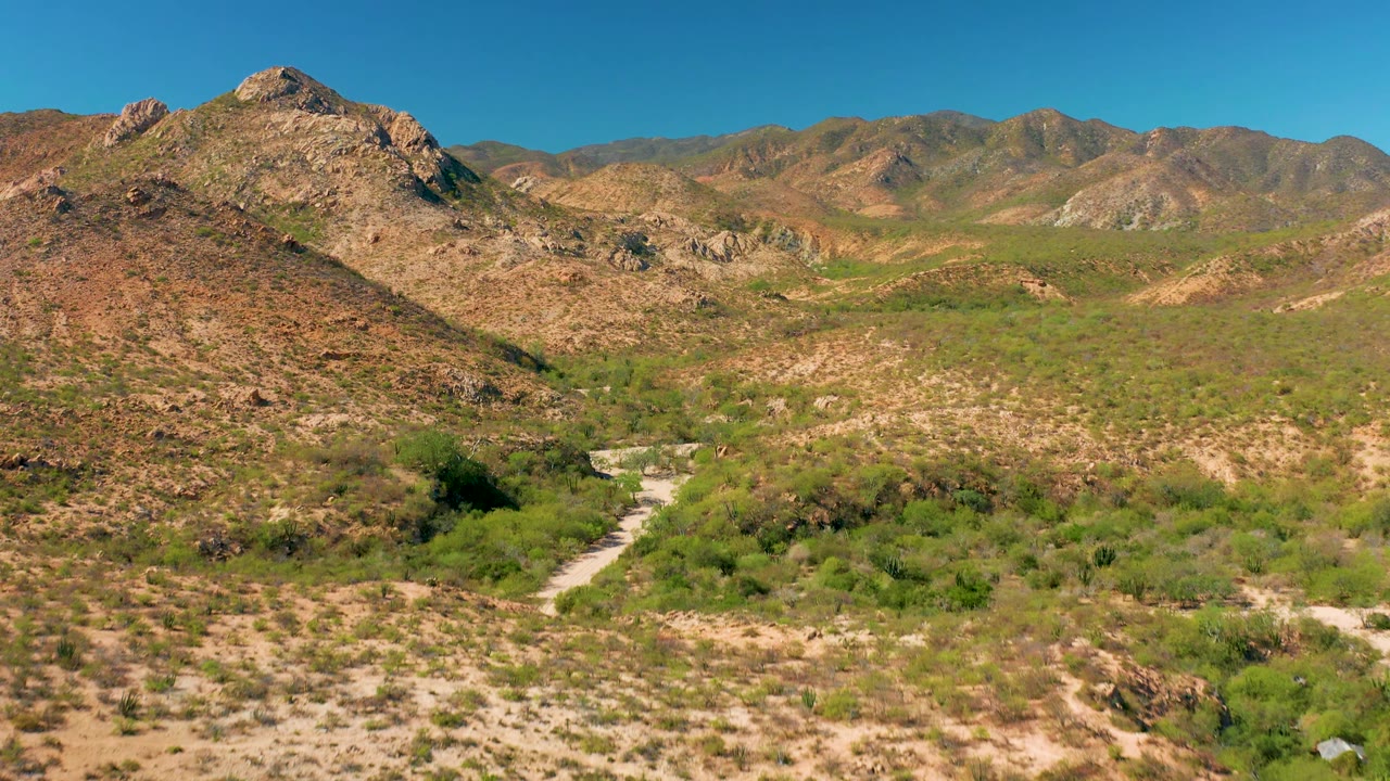 Cabo Pulmo aerial view with the rocky hill dominating the left and center, a bright dirt road curving through scrub in the lower right, layered mountain ridges in back, and no visible people.