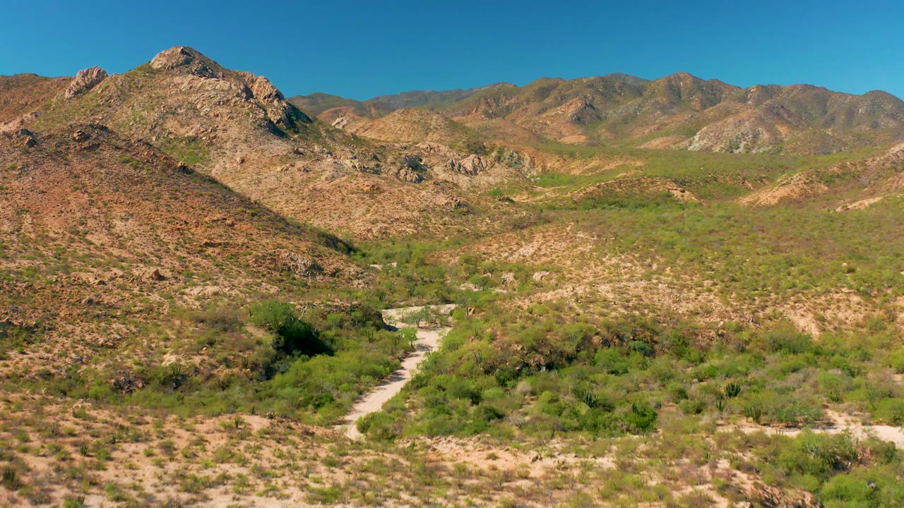 Aerial frame over Cabo Pulmo desert hills with the road entering from the lower left, green shrub patches in the valley, rocky slopes across the middle distance, mountains on the horizon, and no visible people.
