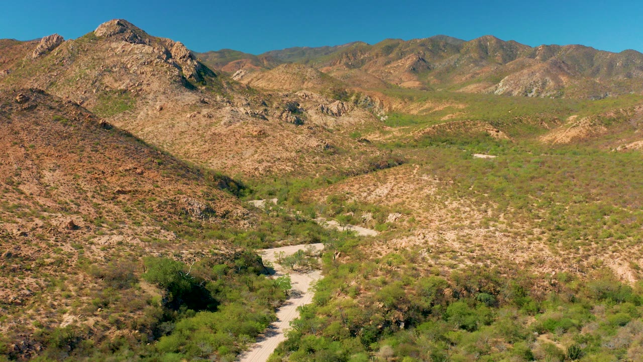 Opening aerial frame over Cabo Pulmo, Baja California Sur, with a pale dirt road crossing a dry valley below rocky hills, distant mountains, blue sky, and no visible people.