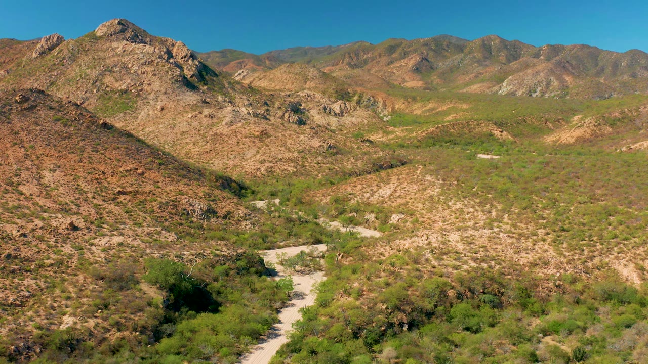 Aerial view in Cabo Pulmo, Baja California Sur, showing the winding dirt road slightly farther into the valley, with rocky brown slopes, green scrub, mountain ridges, blue sky, and no visible people.