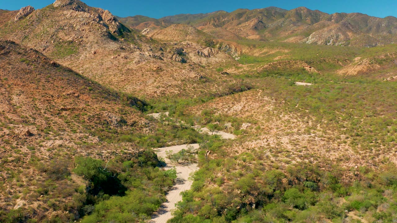 Mid-clip aerial view over Cabo Pulmo, Baja California Sur, where the dirt road curves near the bottom edge beneath rocky hills, sparse green brush, distant mountain slopes, clear sky, and no visible people.