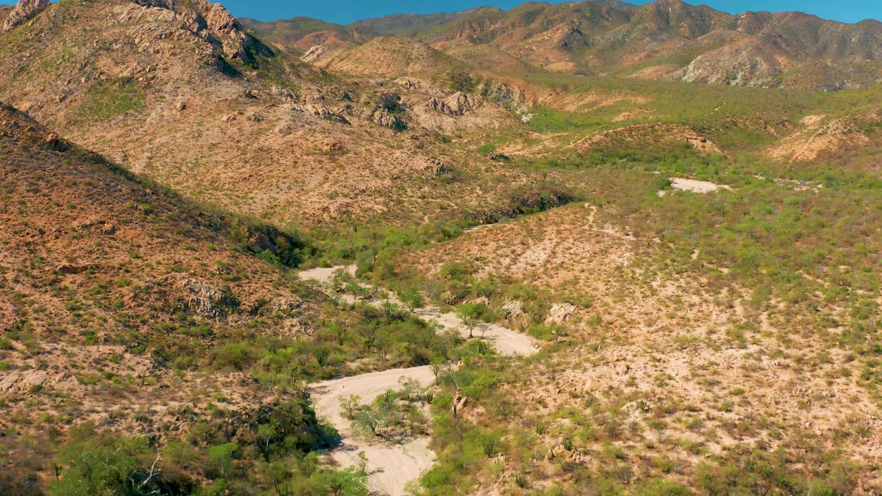 Wide aerial frame of Cabo Pulmo scrubland, a pale riverbed bend, brown rock slopes, distant ridges, and no visible people.