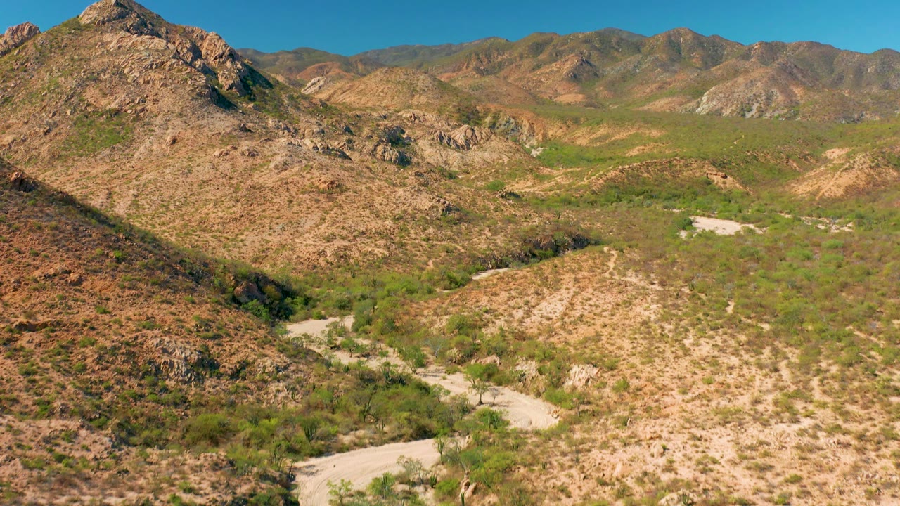 Wide Cabo Pulmo aerial frame with a rocky hill in the foreground, pale wash segments below, layered desert mountains, and no visible people.