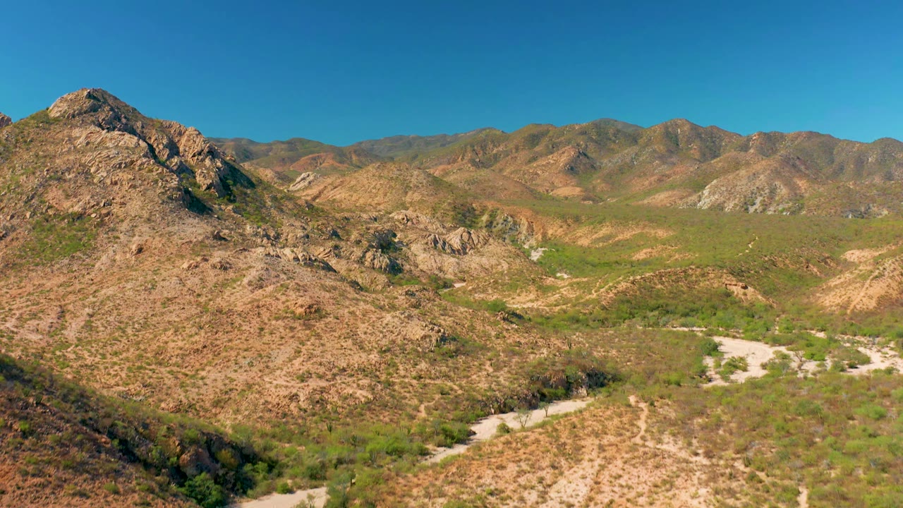 Aerial frame in Cabo Pulmo showing tan hills, scattered green scrub, pale dirt channels through the valley floor, distant mountain ridges, and no visible people.