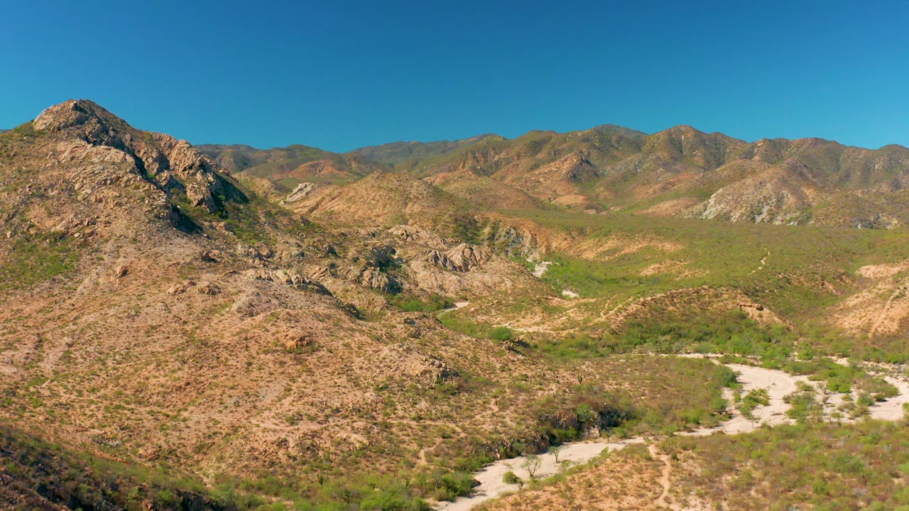 Aerial Cabo Pulmo frame with the rocky foreground slope filling the lower view, a green-brown valley beyond, pale dirt traces, distant mountains, and no visible people.