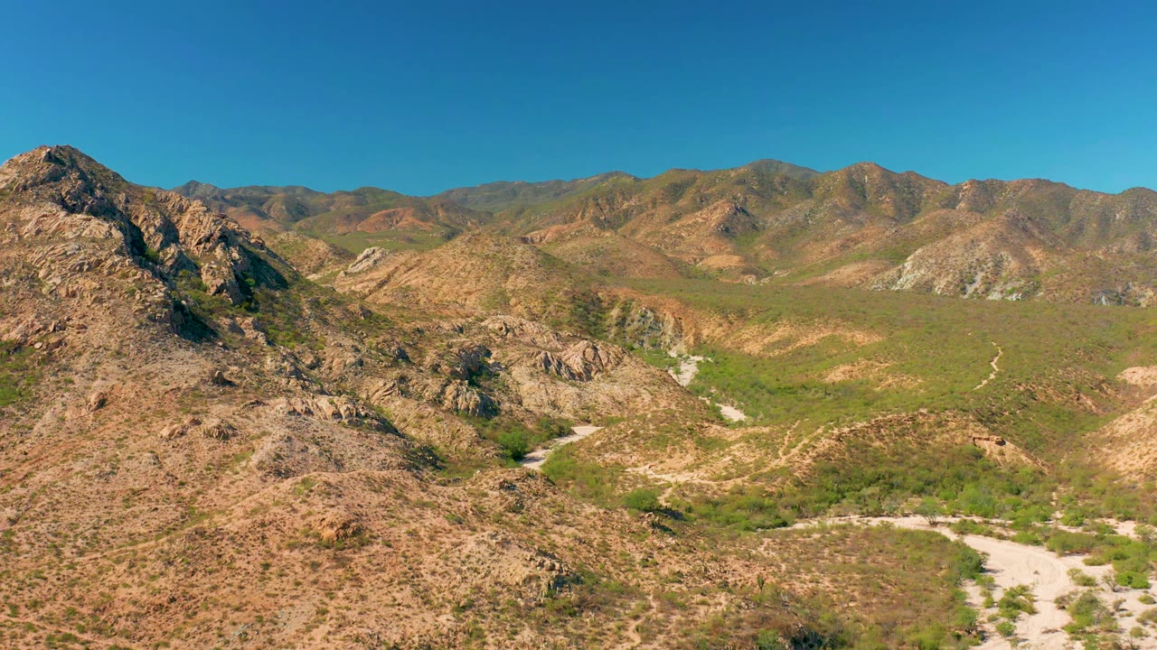 Aerial frame in Cabo Pulmo showing a closer rocky ridge at left, scrub-covered valley floor, pale dirt road bends, distant brown-green mountains, and no visible people.