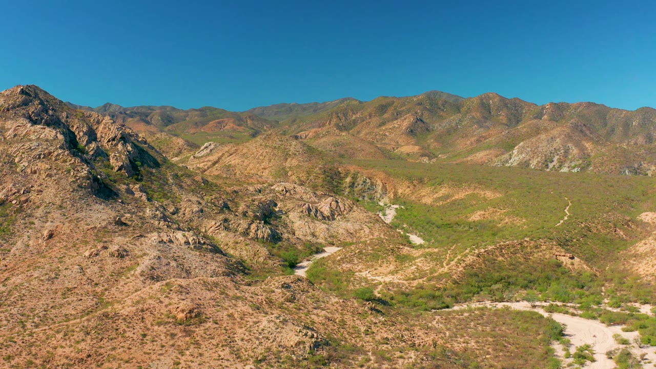 Cabo Pulmo aerial view with the rocky foreground ridge shifted right, a broad green-brown valley, pale road traces through the basin, distant mountains, and no visible people.