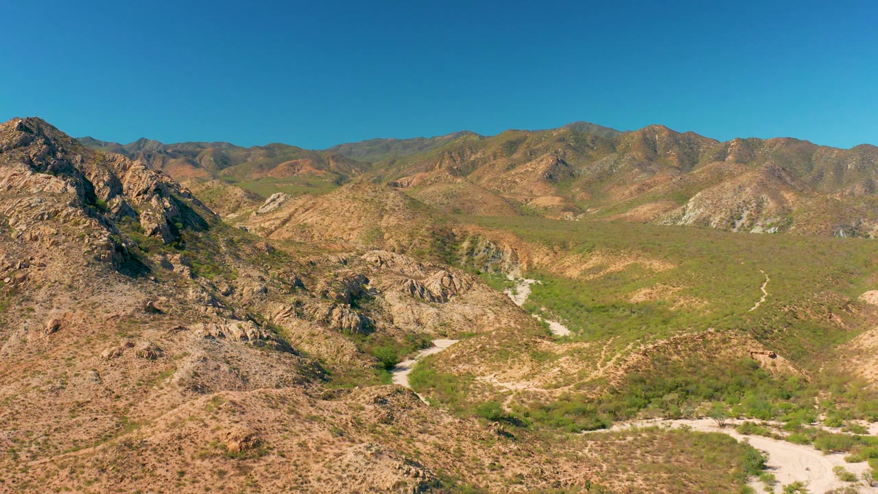 Mid-clip aerial frame over Cabo Pulmo with rocky hills on the left, green scrub across the valley, light dirt road curves near the lower frame, and no visible people.