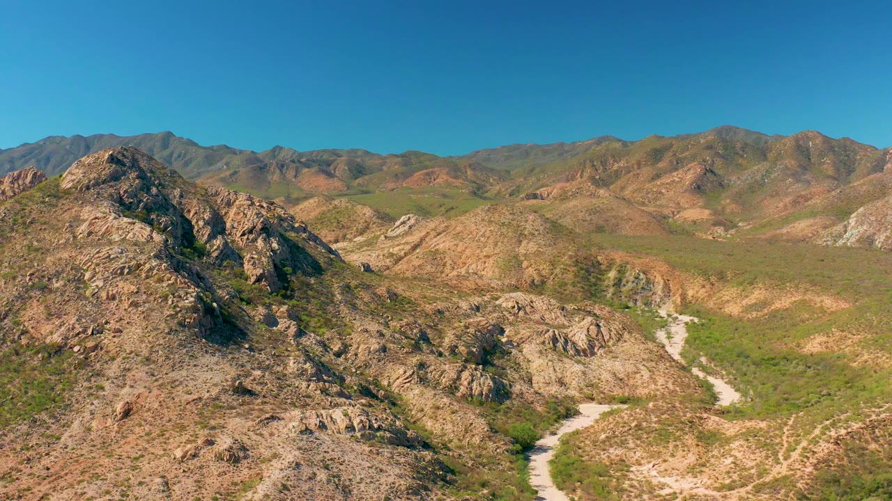Second frame in Cabo Pulmo holds on the same high aerial angle, with the road curving from the lower valley toward rugged ridges and no visible people.