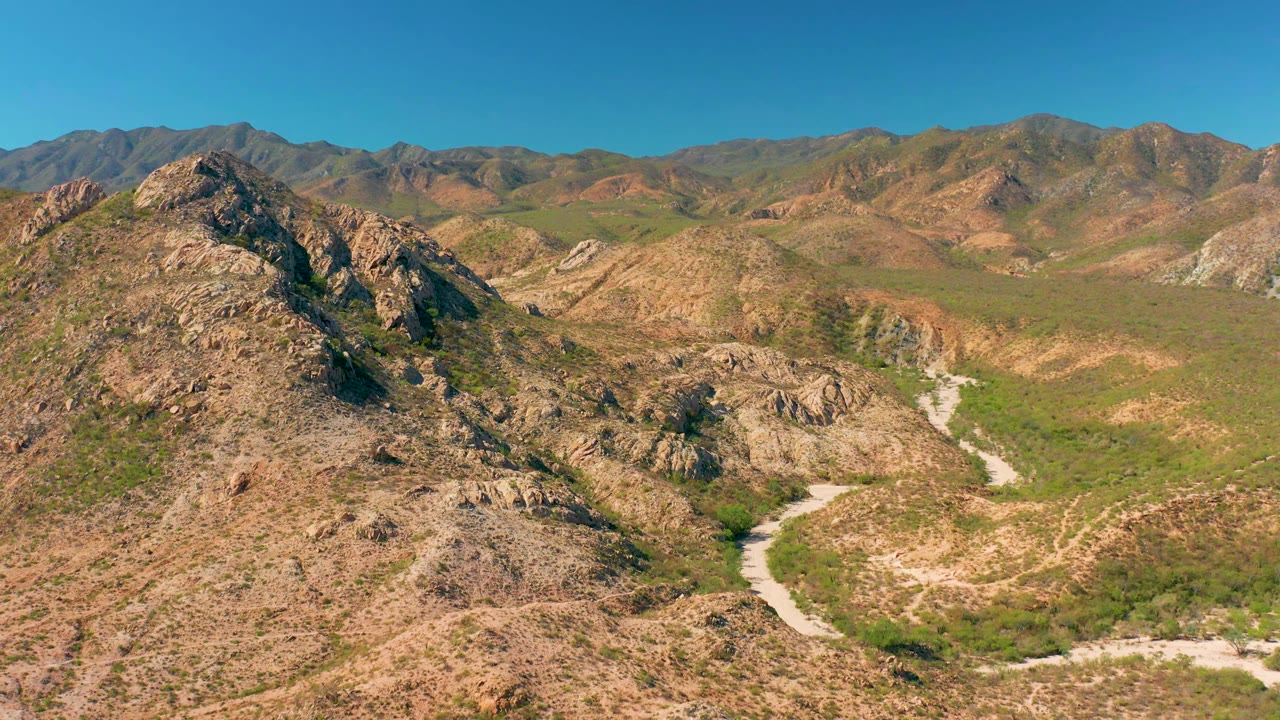 Aerial view in Cabo Pulmo with a rocky hill filling the foreground, the dirt road curving through the valley, distant mountains, and no visible people.