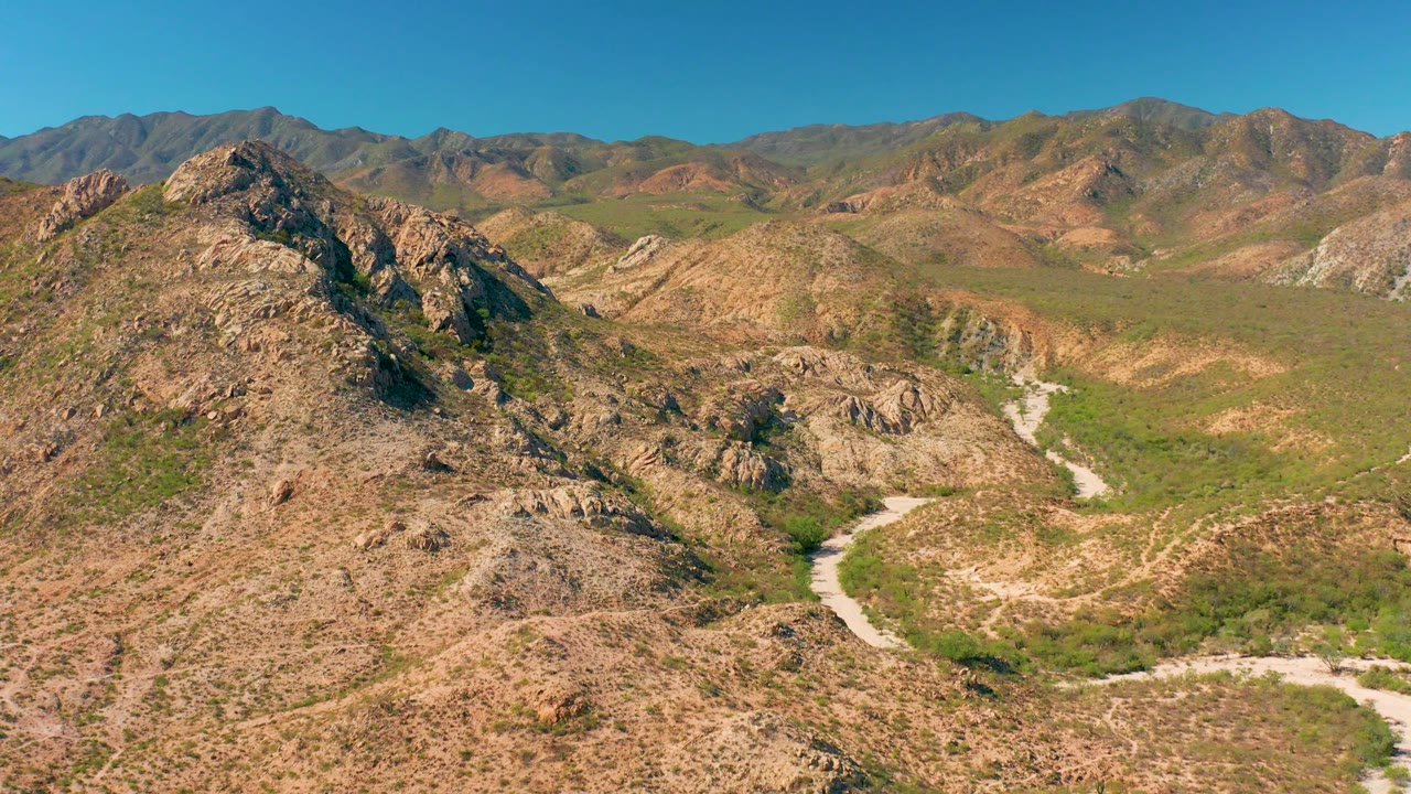 Aerial Cabo Pulmo landscape with a rocky hill at left, a winding dirt road below, scrubby green vegetation, layered mountain ridges, and no visible people.