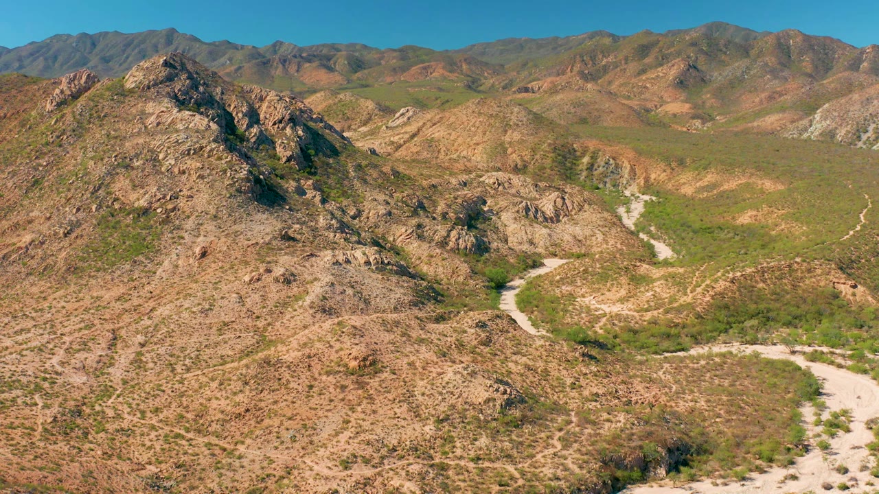 Aerial frame in Cabo Pulmo showing the rocky central hill more prominently, with the pale winding road crossing the lower valley, green scrub, distant mountains, and no visible people.